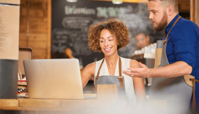 Imagem faz referência à pequenas empresas, em que um casal com avental está em frente a um computador analisando o planejamento financeiro do negócio.